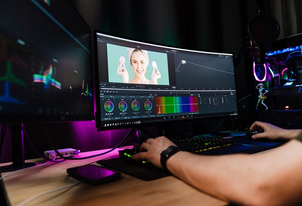 Back view of a young white man video editor sitting in front of a big screen working on a video montage indoors